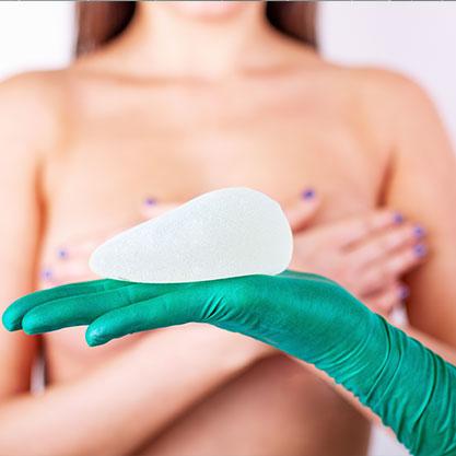 Steriler Handschuh auf dem eine Silikonprothese liegt vor einer jungen, unbekleideten Frau.  Dieses Bild demonstriert die Möglichkeit einer Brustvergrößerung mit Silikonprothesen